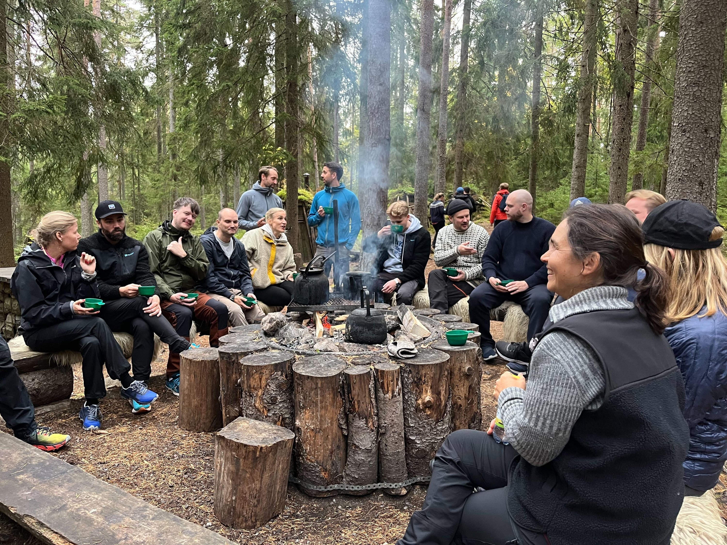 En grupp människor sitter runt en lägereld i en skog, njuter av fika och samtal i en avkopplande utomhusmiljö