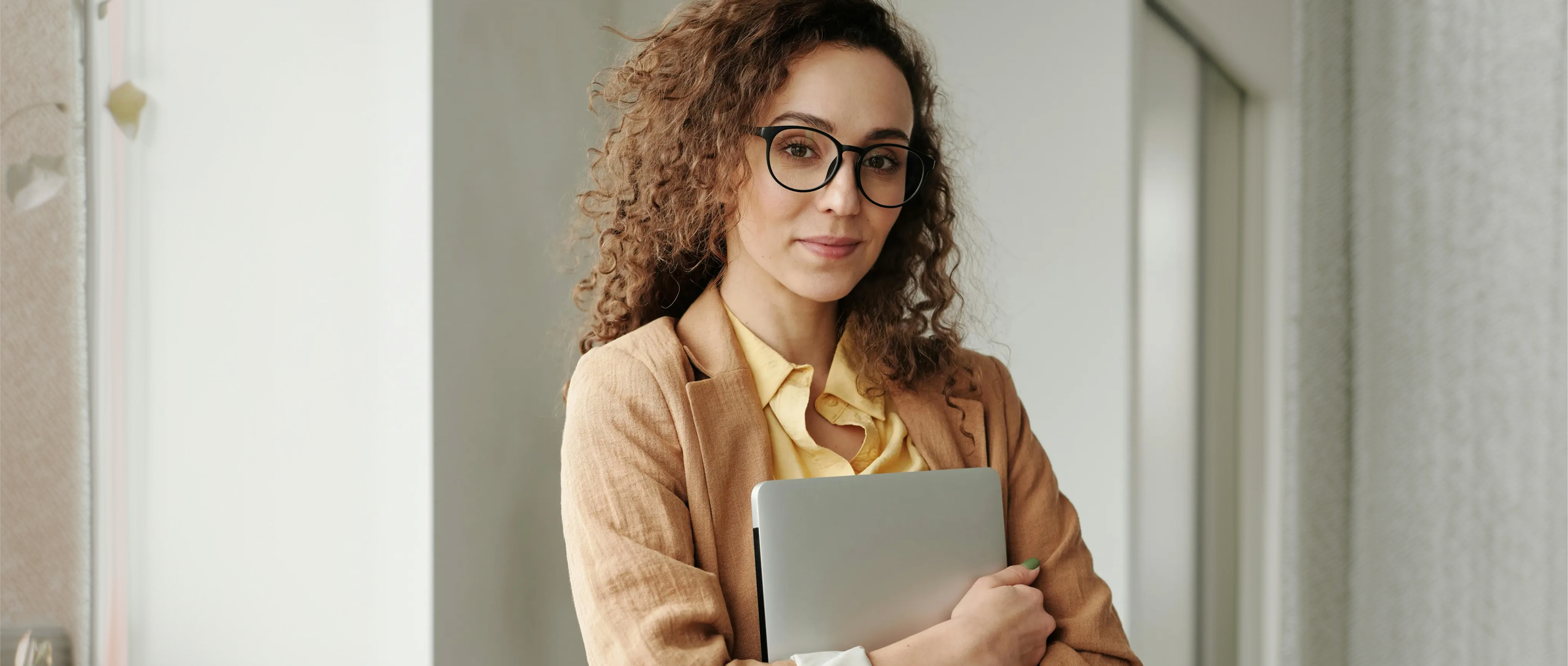 En kvinna med brunt lockigt hår och glasögon ler och håller i en laptop